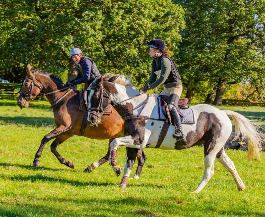 Two horse riders cantering in a field, wearing a Beige Equiseat Aid
